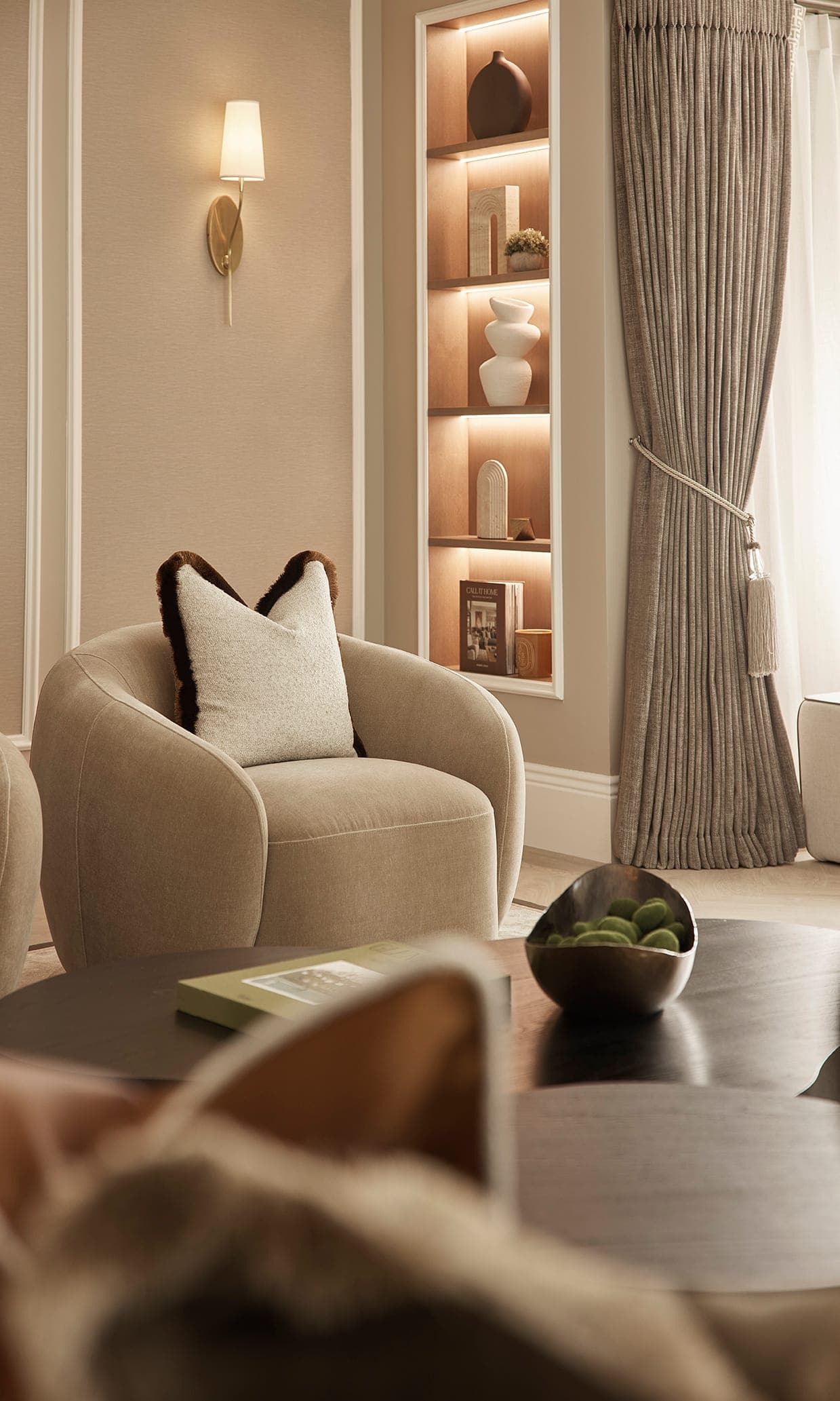 Curved armchair with a fringed cushion beside lit alcove shelving in the family room of a Prestbury home